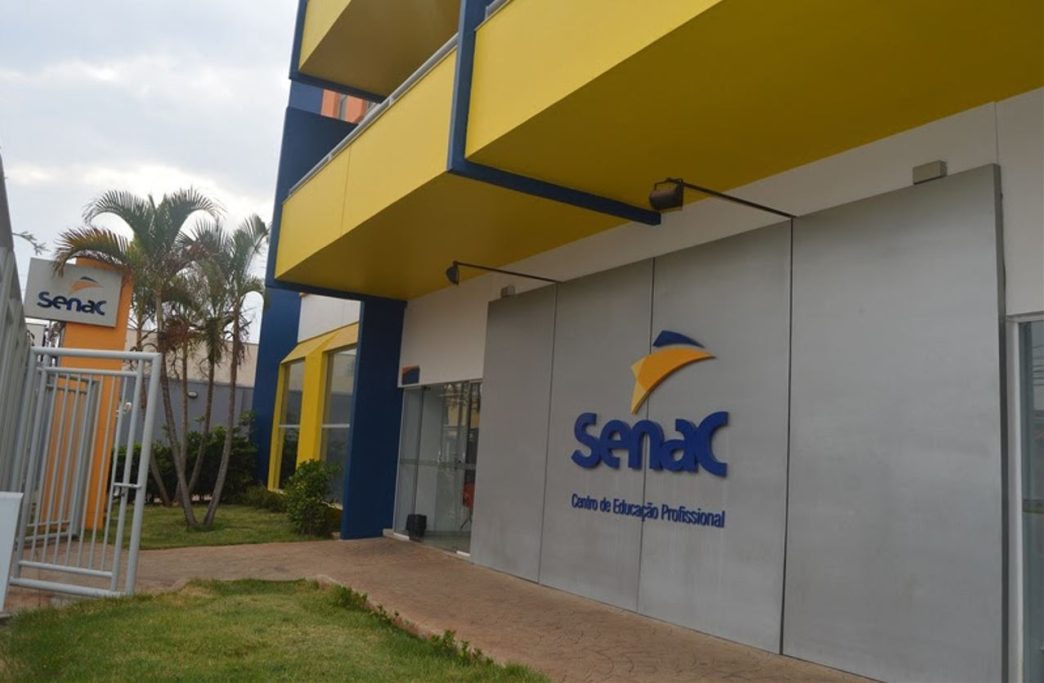Fachada moderna do Senac Uberlândia, com estrutura em tons de azul e amarelo, jardins bem cuidados e o logotipo da instituição em destaque na entrada principal.