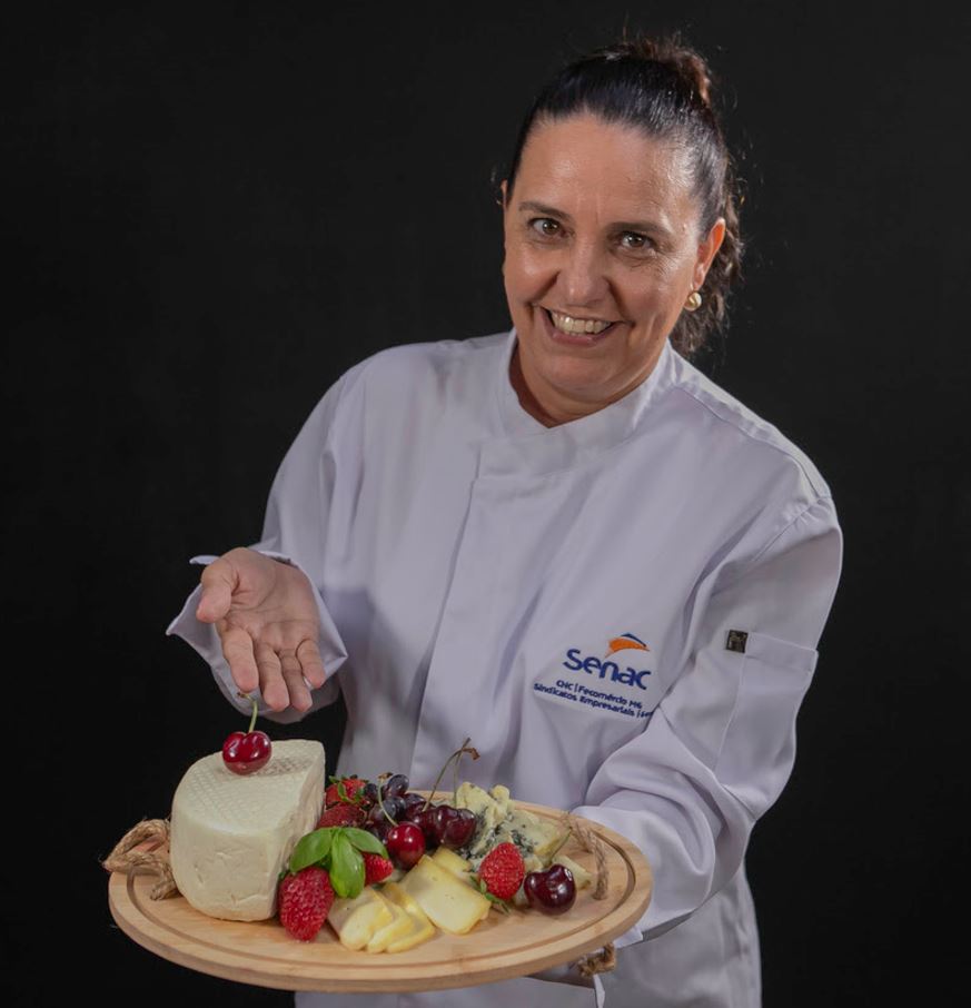 Agnes Laila, especialista em gastronomia e sommelière do Senac, sorri ao apresentar uma tábua de queijos e frutas frescas, usando uniforme branco com o logotipo do Senac, sobre fundo escuro.