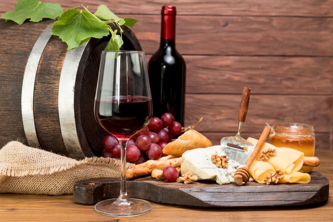 Tábua rústica com queijos variados, uvas, nozes, pão e mel, acompanhada por taça e garrafa de vinho tinto, em frente a um barril de madeira.
