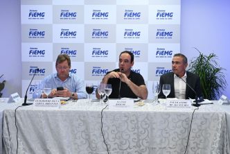 Três representantes de entidades industriais e jornalísticas sentados à mesa durante coletiva de imprensa do balanço econômico da FIEMG, com logotipos da FIEMG ao fundo.