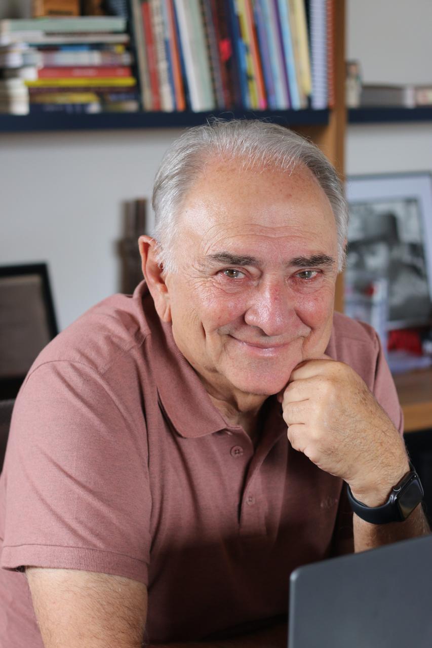 Retrato do escritor Luís Giffoni, sorrindo e apoiando o queixo na mão, em ambiente de escritório com estante de livros ao fundo.