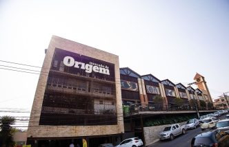 Fachada do Mercado de Origem, em Belo Horizonte, com arquitetura moderna, torre com relógio e fila de carros estacionados.