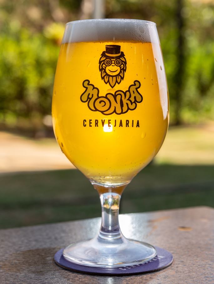 Taça personalizada da Monka Cervejaria, repleta de cerveja dourada com espuma cremosa, sobre mesa externa iluminada pelo sol.