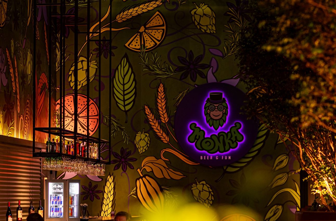 Interior da Monka Cervejaria em Belo Horizonte, com parede ilustrada por elementos botânicos e o logotipo iluminado da marca ao centro, transmitindo um ambiente acolhedor e moderno com iluminação âmbar e decoração temática.