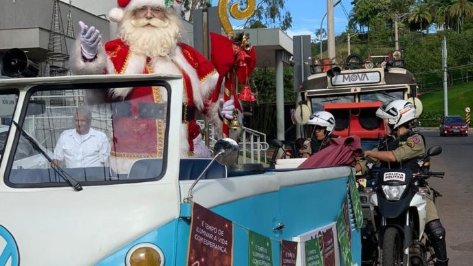 Papai Noel acena sorridente em desfile natalino a bordo de uma Kombi decorada no Natal Solidário Vila da Serra, acompanhado por policiais militares e personagens natalinos.