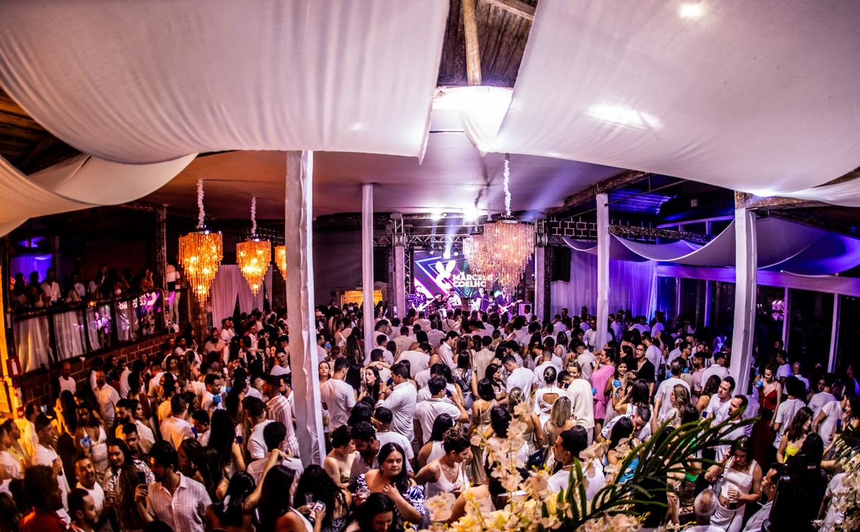 Grande salão decorado com tecidos brancos e lustres de cristal, lotado de pessoas vestidas de branco celebrando o Réveillon ao som de banda ao vivo, sob luzes roxas e ambiente elegante.
