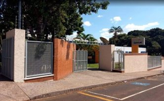 Imagem da fachada principal do Senac em Araxá, com portão de entrada aberto, muros em alvenaria, vegetação ao fundo e o letreiro institucional visível sob céu azul com nuvens claras.