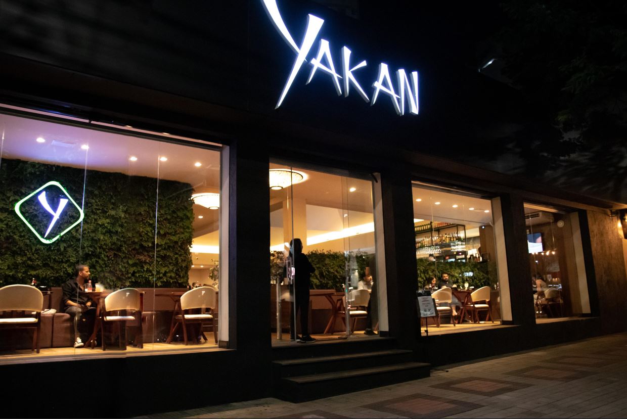 Fachada noturna iluminada do restaurante japonês Yakan, com amplas janelas de vidro revelando o ambiente interno moderno, com clientes sendo atendidos em mesas sofisticadas. Letreiro luminoso com o nome "YAKAN" destacado acima da entrada.