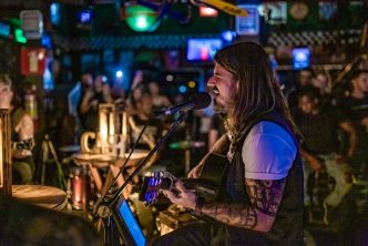 Músico da banda Cash se apresenta ao vivo em ambiente intimista, tocando violão e cantando em um palco iluminado, rodeado pelo público.