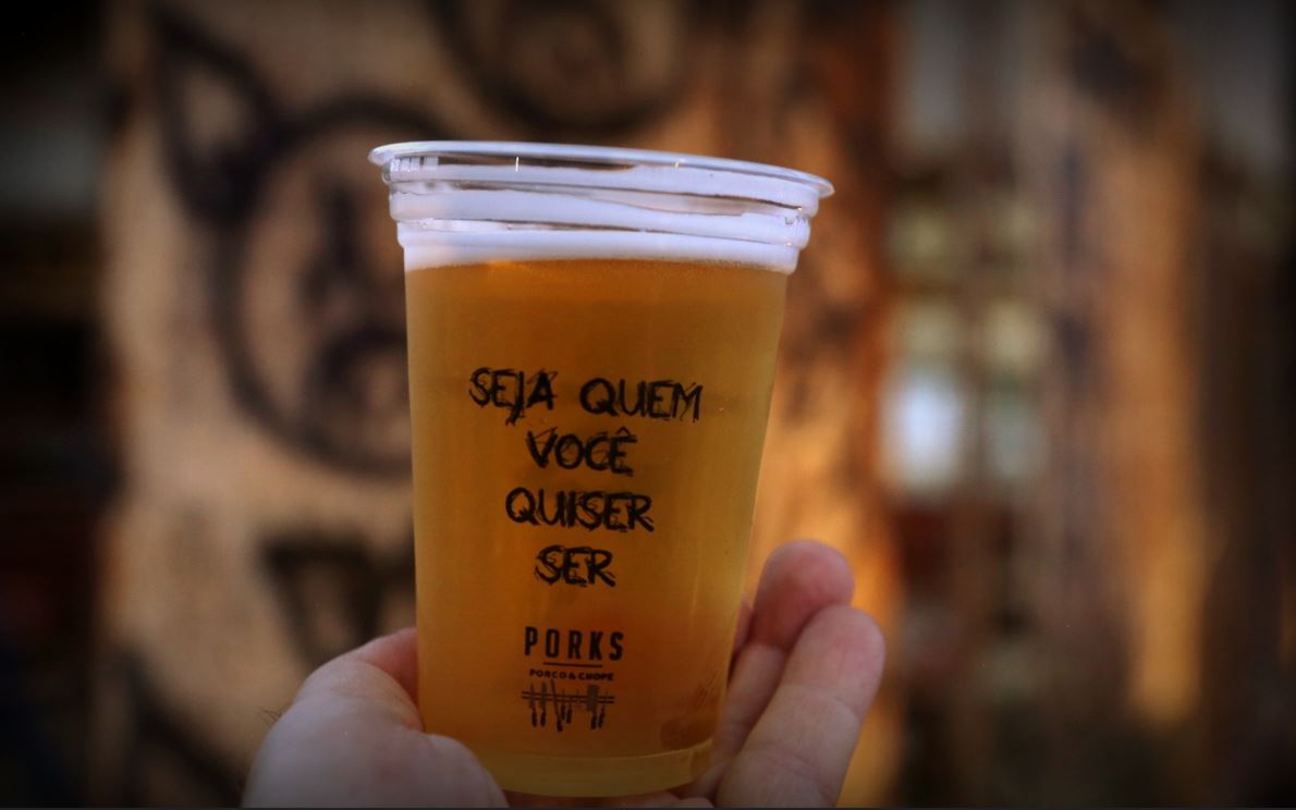 Copo de chope claro e gelado com a frase "Seja quem você quiser ser" estampada, segurado por uma mão em frente a um fundo com arte urbana desfocada, no bar Porks.