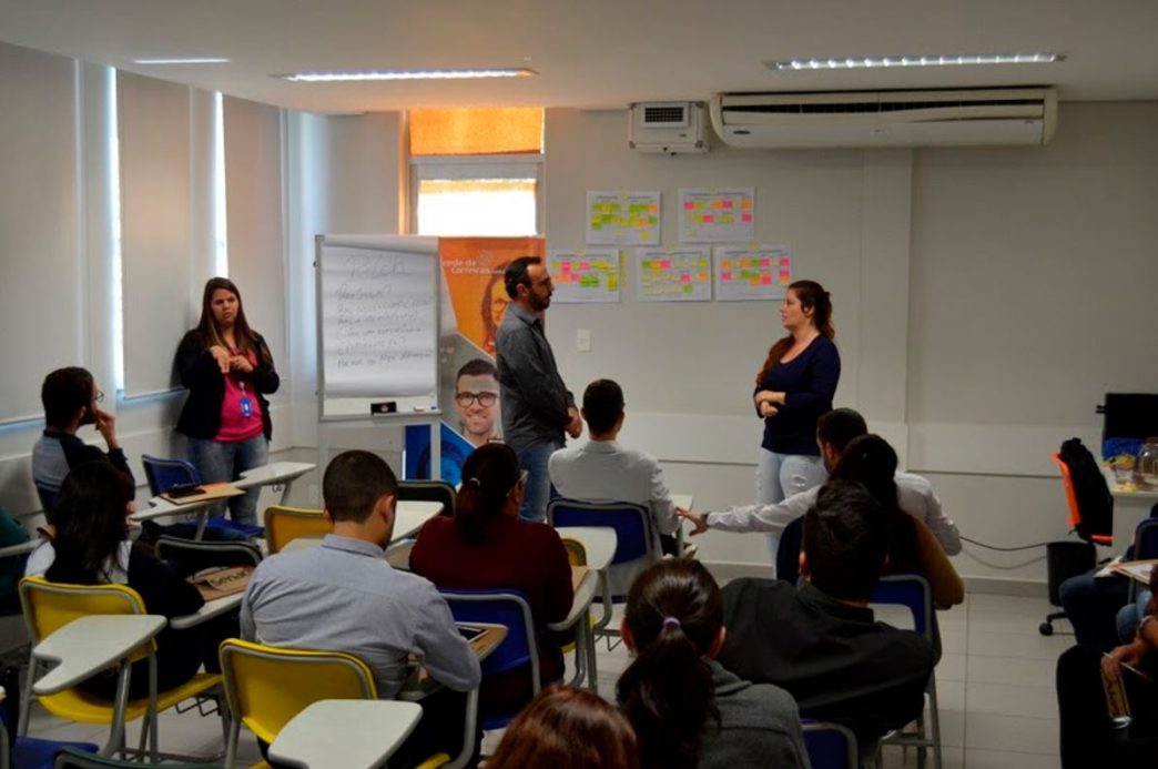 Sala de aula com alunos participando de atividade prática durante curso profissionalizante; três instrutores conduzem dinâmica interativa.