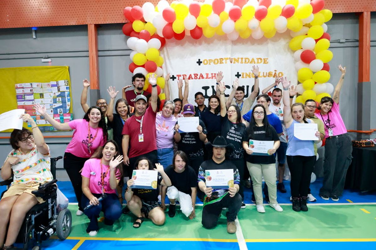 Grupo diverso de formandos, incluindo jovens com deficiência intelectual e mães empreendedoras, celebrando a conclusão de cursos profissionalizantes promovidos pela parceria entre o Senac Minas e o Instituto Mano Down, diante de um painel decorado com balões coloridos e mensagens de inclusão, trabalho e oportunidade.