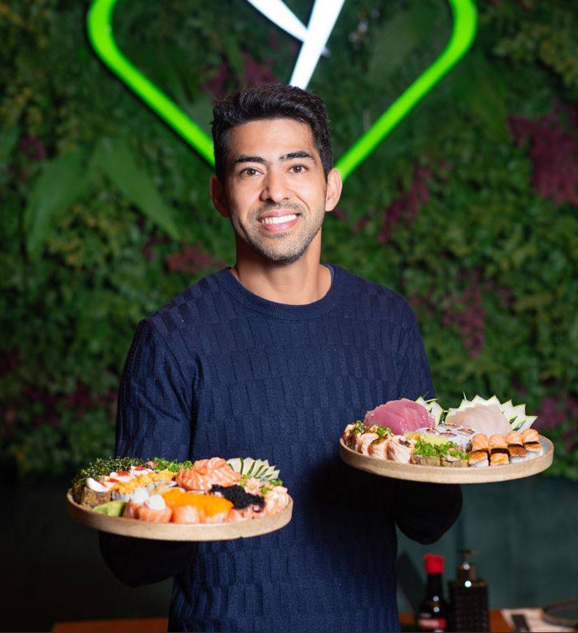 Homem sorridente segura duas tábuas com variedade de sushis e sashimis no restaurante Yakan, diante de uma parede verde com o logo iluminado da casa.