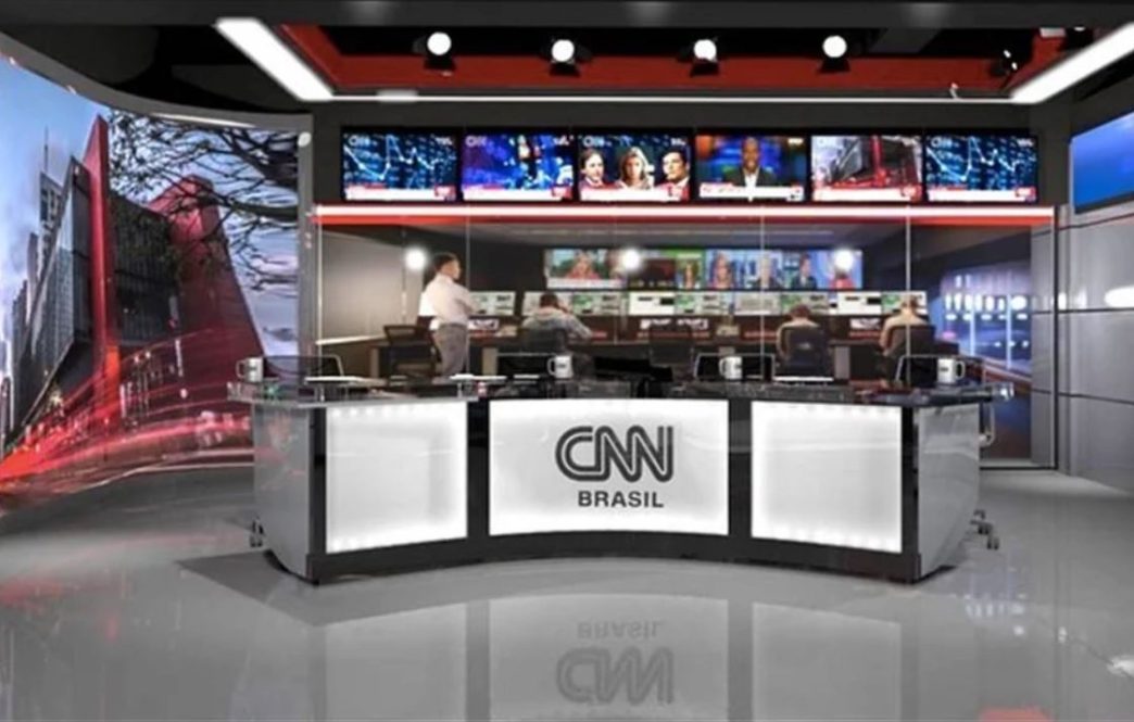 Estúdio da CNN Brasil com bancada de jornalismo em primeiro plano, telões ao fundo exibindo programação ao vivo e equipe técnica operando nos bastidores.