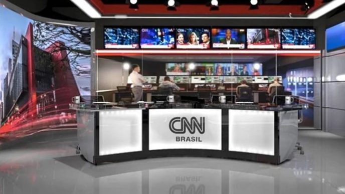 Estúdio da CNN Brasil com bancada de jornalismo em primeiro plano, telões ao fundo exibindo programação ao vivo e equipe técnica operando nos bastidores.
