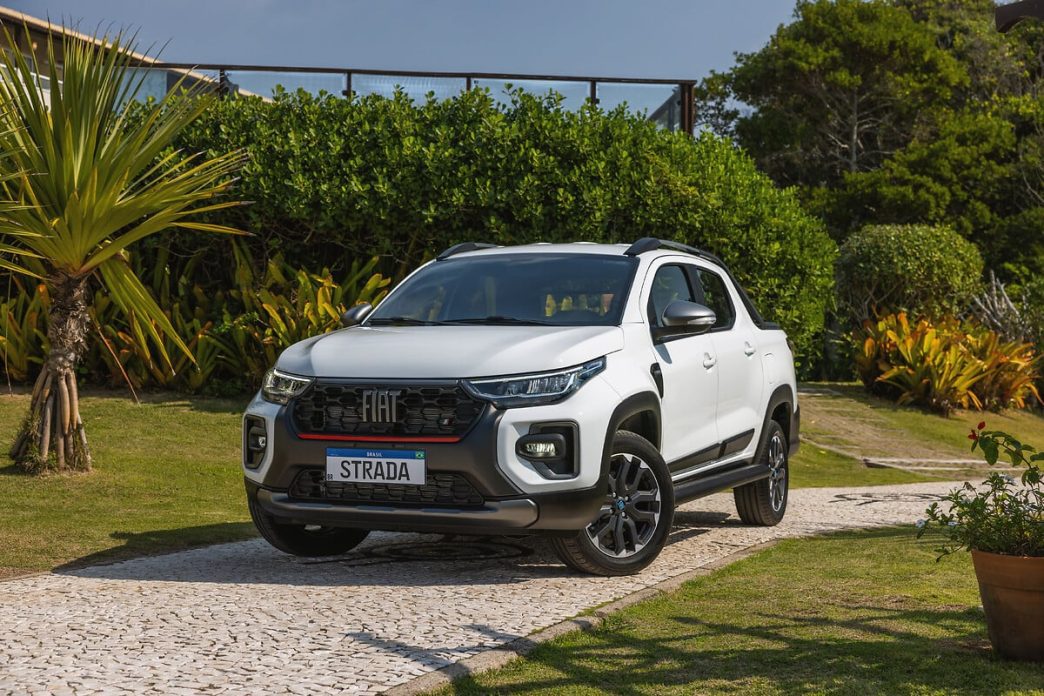 Fiat Strada 2025 branca, versão cabine dupla, estacionada em jardim tropical com fundo arborizado, destacando design robusto.