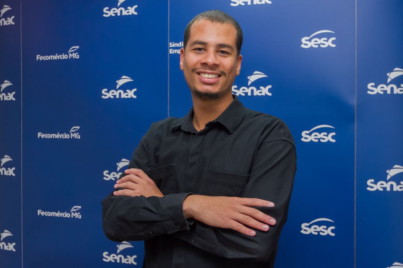 Guilherme Monteiro, Analista de Empregabilidade do Senac em Minas, posando sorridente com os braços cruzados diante de painel institucional.