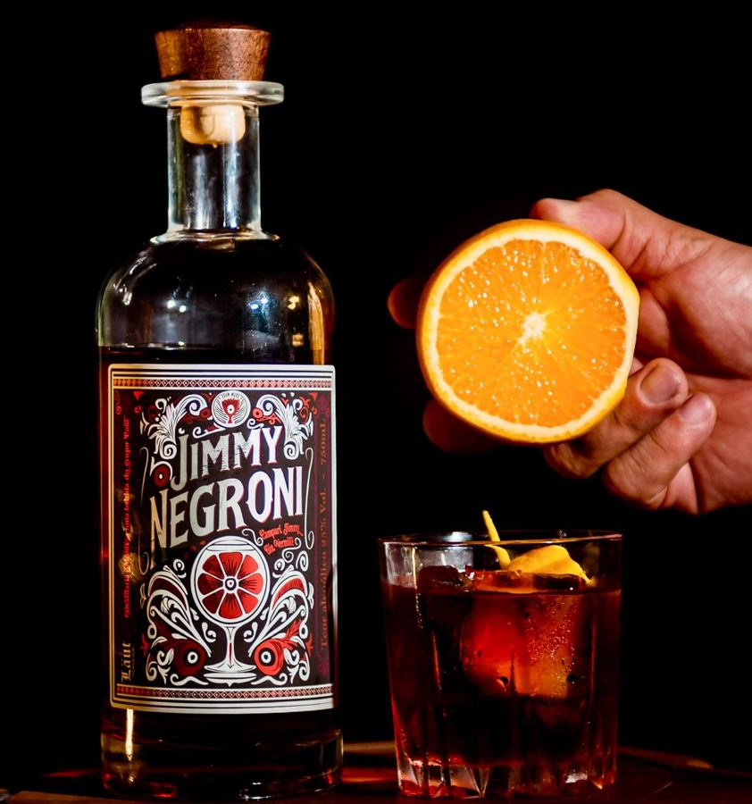 Garrafa de Jimmy Negroni ao lado de um copo com o drinque servido com gelo e casca de laranja, e uma mão segurando meia laranja fresca.