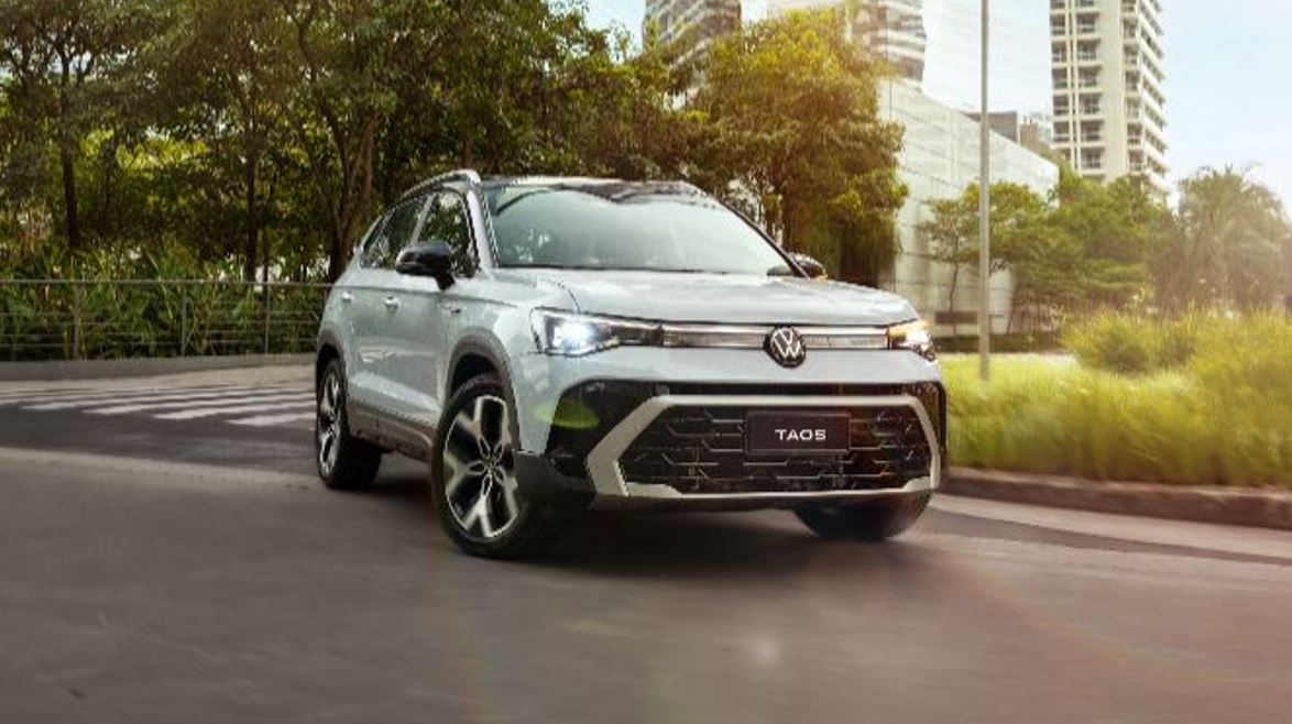 Volkswagen Taos 2026 branco circulando em área urbana arborizada, com destaque para o novo design frontal e luzes diurnas em LED.