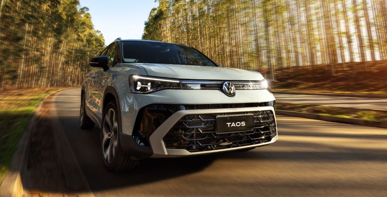 Volkswagen Taos 2026 em movimento por estrada sinuosa cercada por árvores, evidenciando grade frontal e assinatura em LED.