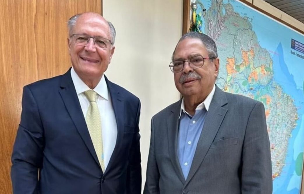 Vice-presidente Geraldo Alckmin ao lado do jornalista Paulo César de Oliveira (PCO), ambos de terno, posando sorridentes em ambiente institucional com mapa do Brasil ao fundo.