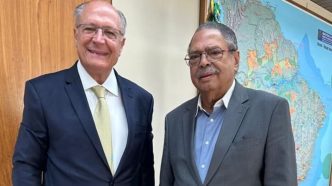Vice-presidente Geraldo Alckmin ao lado do jornalista Paulo César de Oliveira (PCO), ambos de terno, posando sorridentes em ambiente institucional com mapa do Brasil ao fundo.