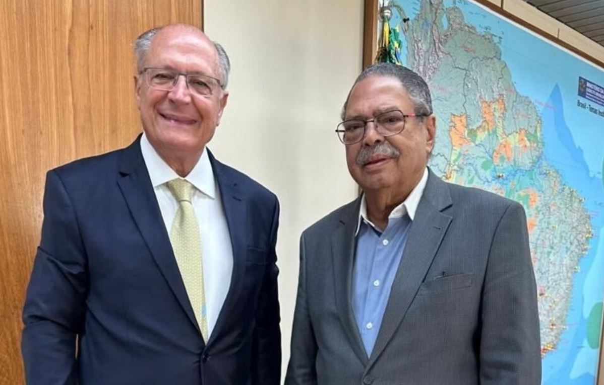 Vice-presidente Geraldo Alckmin ao lado do jornalista Paulo César de Oliveira (PCO), ambos de terno, posando sorridentes em ambiente institucional com mapa do Brasil ao fundo.