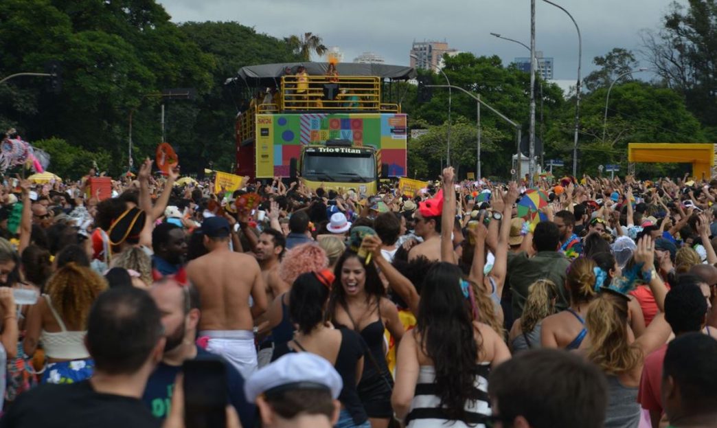 Multidão de foliões acompanha trio elétrico colorido em desfile de bloco de Carnaval de rua, com fantasias e clima de festa popular.