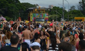 Multidão de foliões acompanha trio elétrico colorido em desfile de bloco de Carnaval de rua, com fantasias e clima de festa popular.
