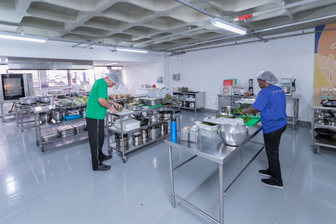 Profissionais utilizando a cozinha pedagógica da Faculdade Senac em Belo Horizonte, equipada com bancadas de inox, utensílios e ingredientes frescos.