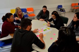 Grupo de alunos e alunas participa de atividade em sala de aula, sentados ao redor de uma mesa redonda, interagindo com post-its coloridos durante dinâmica colaborativa de curso gratuito promovido pelo Senac em Caeté.