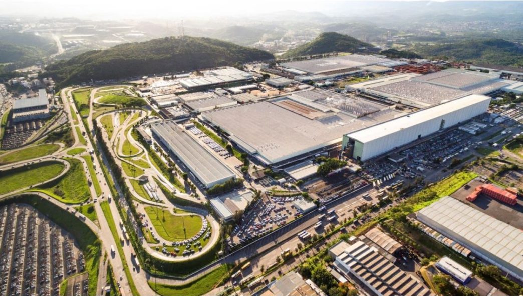 Vista aérea do Polo Automotivo Stellantis em Betim, Minas Gerais, com instalações industriais, pistas de testes e área logística cercadas por vegetação e montanhas ao fundo.