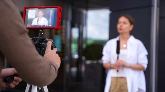 Profissional de multimídia sendo gravada durante entrevista externa, com cinegrafista operando câmera e monitor de vídeo.