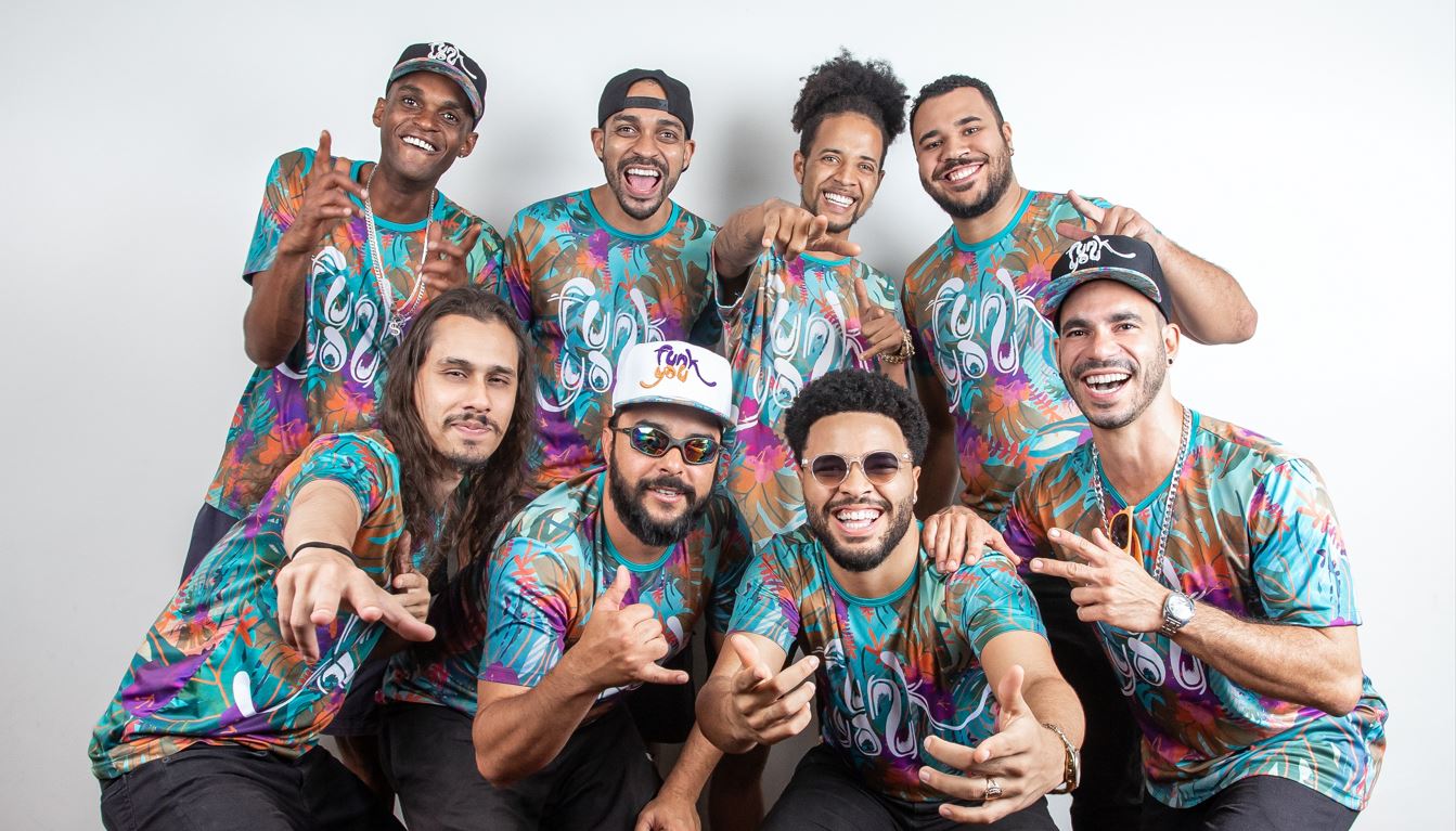 Integrantes da banda Funk You posam sorrindo com camisetas coloridas em estúdio, destacando a energia do Carnaval de BH.