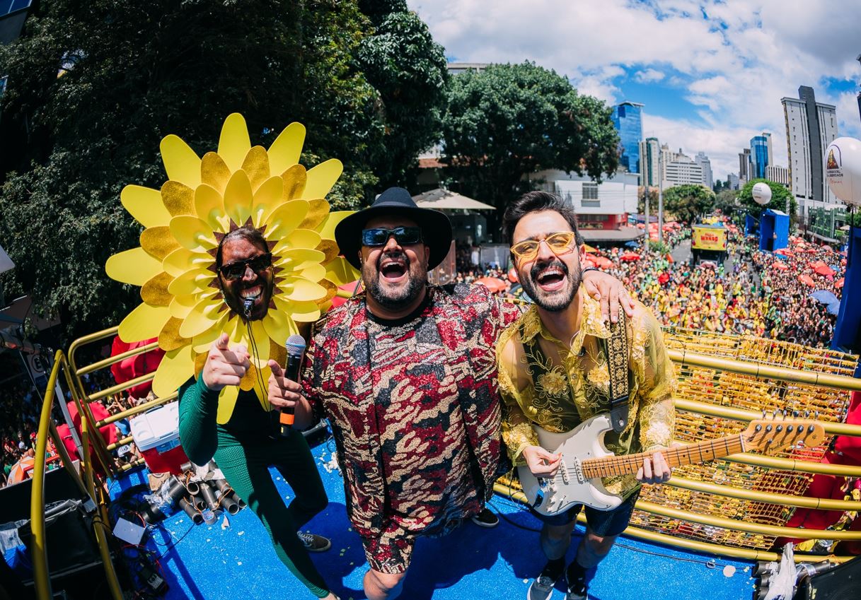 Três integrantes do Bloco Beiço do Wando animam foliões sobre um trio elétrico em Belo Horizonte, com fantasias coloridas e expressões alegres, diante de uma multidão vestida com roupas carnavalescas nas ruas do centro da cidade sob o céu azul.