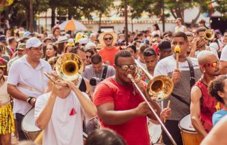 Banda de metais anima multidão durante Carnaval de rua em Minas Gerais, com músicos tocando trombone e percussão entre foliões.