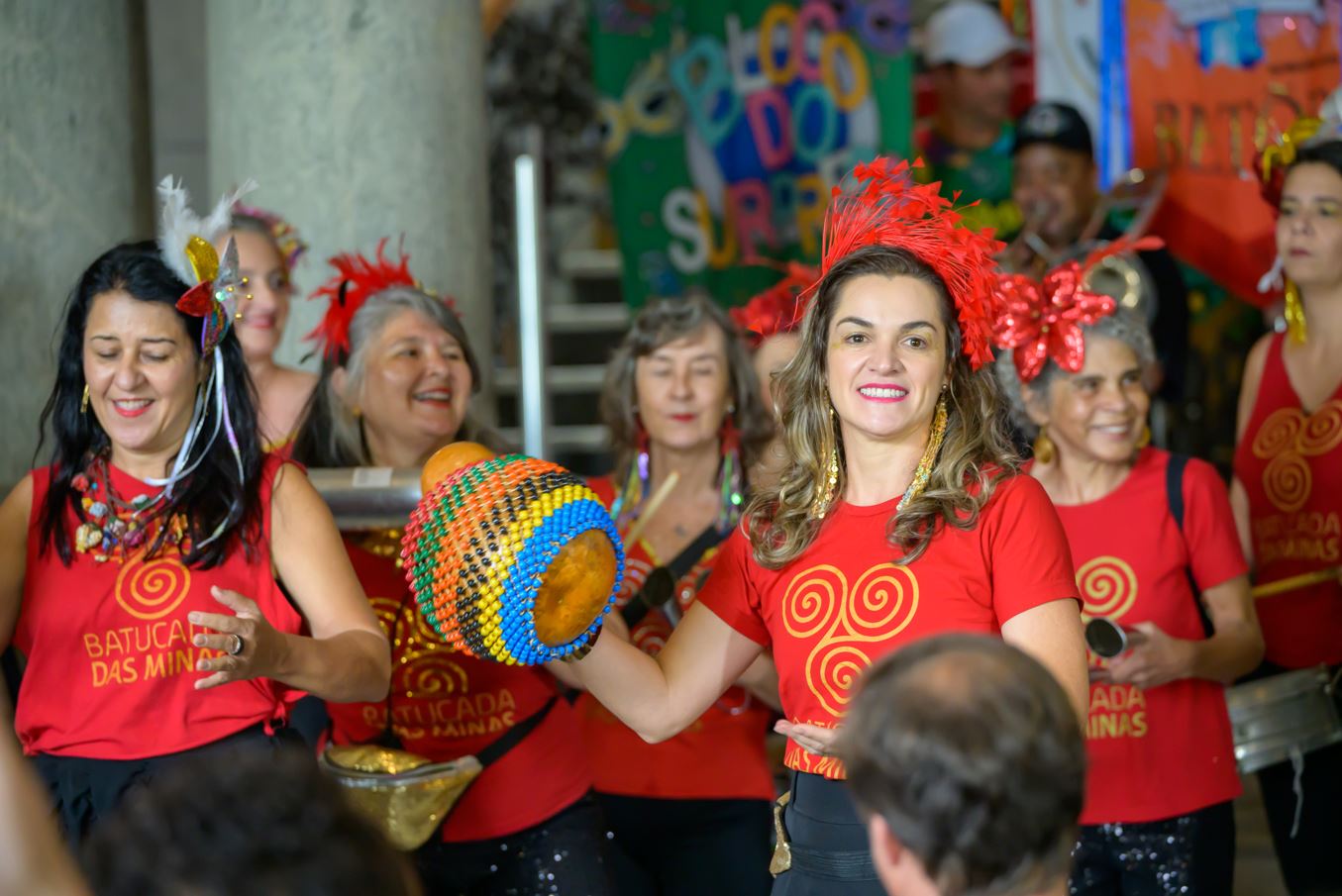 Mulheres integrantes de bloco carnavalesco se apresentam com instrumentos de percussão durante o Carnaval nas Cidades Históricas de Minas Gerais, usando camisetas vermelhas e adereços coloridos, em ambiente festivo e cultural.