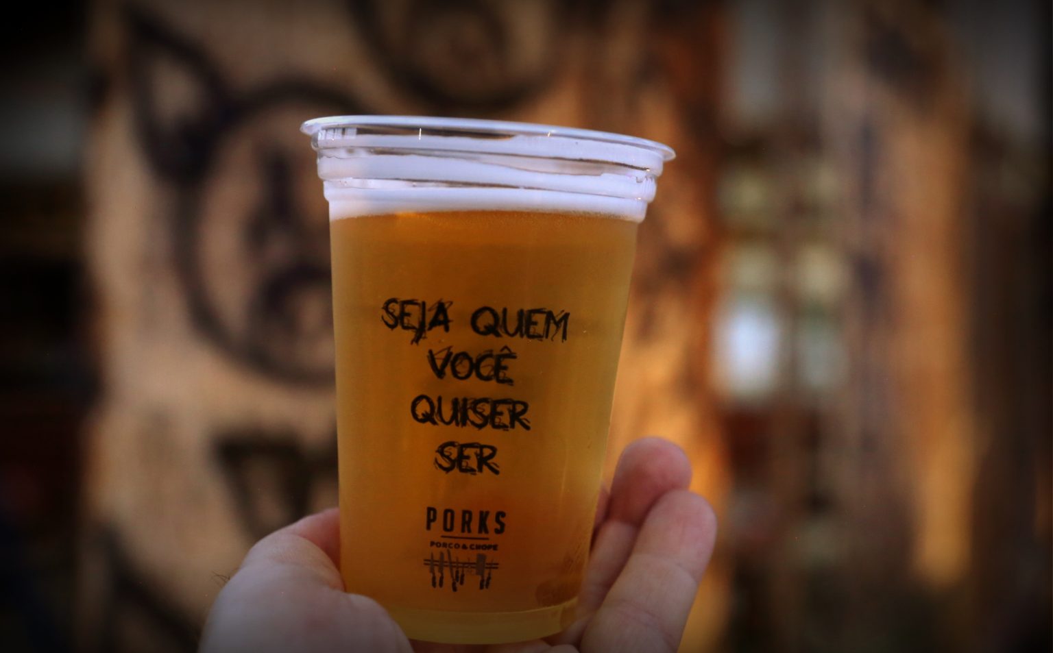 Copo de chope do bar Porks, com a frase "Seja quem você quiser ser", sendo segurado por uma mão na Praça Tiradentes.