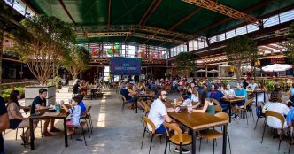 Público reunido no Mercado de Origem, em Belo Horizonte, em ambiente coberto e arejado, com mesas ocupadas por famílias e amigos aproveitando momentos de lazer, gastronomia e convivência.