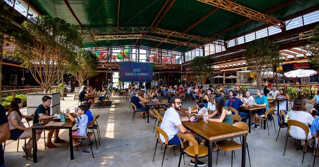 Público reunido no Mercado de Origem, em Belo Horizonte, em ambiente coberto e arejado, com mesas ocupadas por famílias e amigos aproveitando momentos de lazer, gastronomia e convivência.