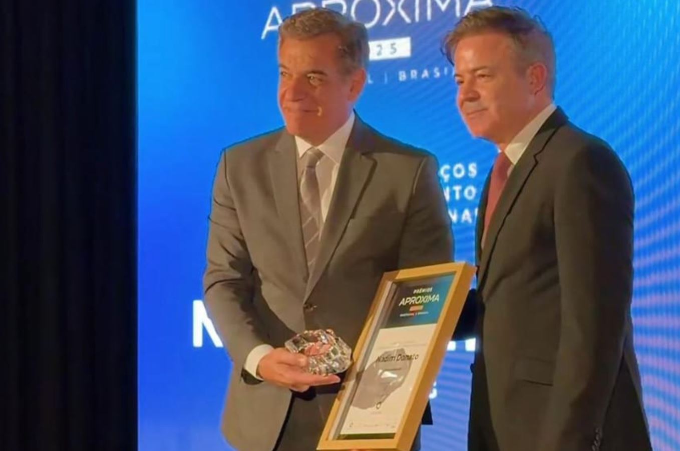 Nadim Elias Donato Filho recebe o Prêmio Aproxima Portugal–Brasil durante cerimônia no Porto, segurando troféu e certificado ao lado de representante da organização.