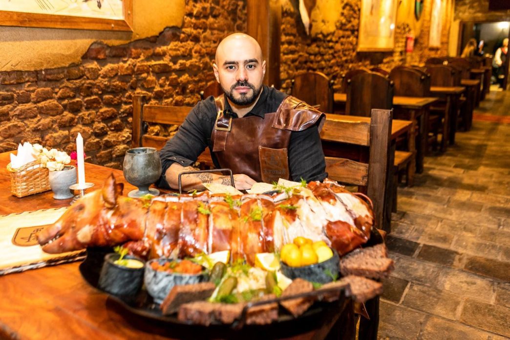 Homem com traje medieval sentado à mesa na Forja Taverna, na Savassi, em Belo Horizonte, diante de um porco assado inteiro servido com legumes, pães rústicos e bebidas em ambiente com decoração temática de taverna.