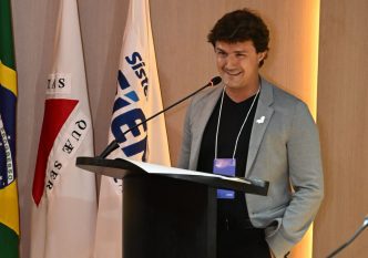 Jovem empresário discursa ao púlpito durante cerimônia do FIEMG Jovem, em Belo Horizonte, com bandeiras institucionais ao fundo.