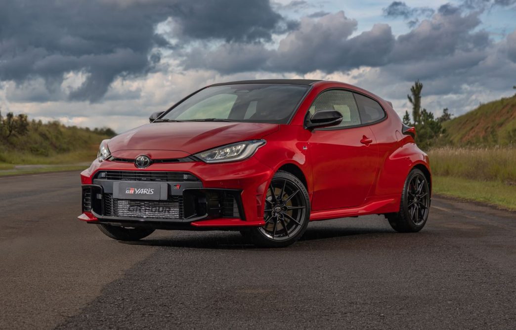 Toyota GR Yaris vermelho estacionado em pista asfaltada, com design esportivo, grade frontal ampla, rodas pretas de 18 polegadas e teto em fibra de carbono sob céu nublado.