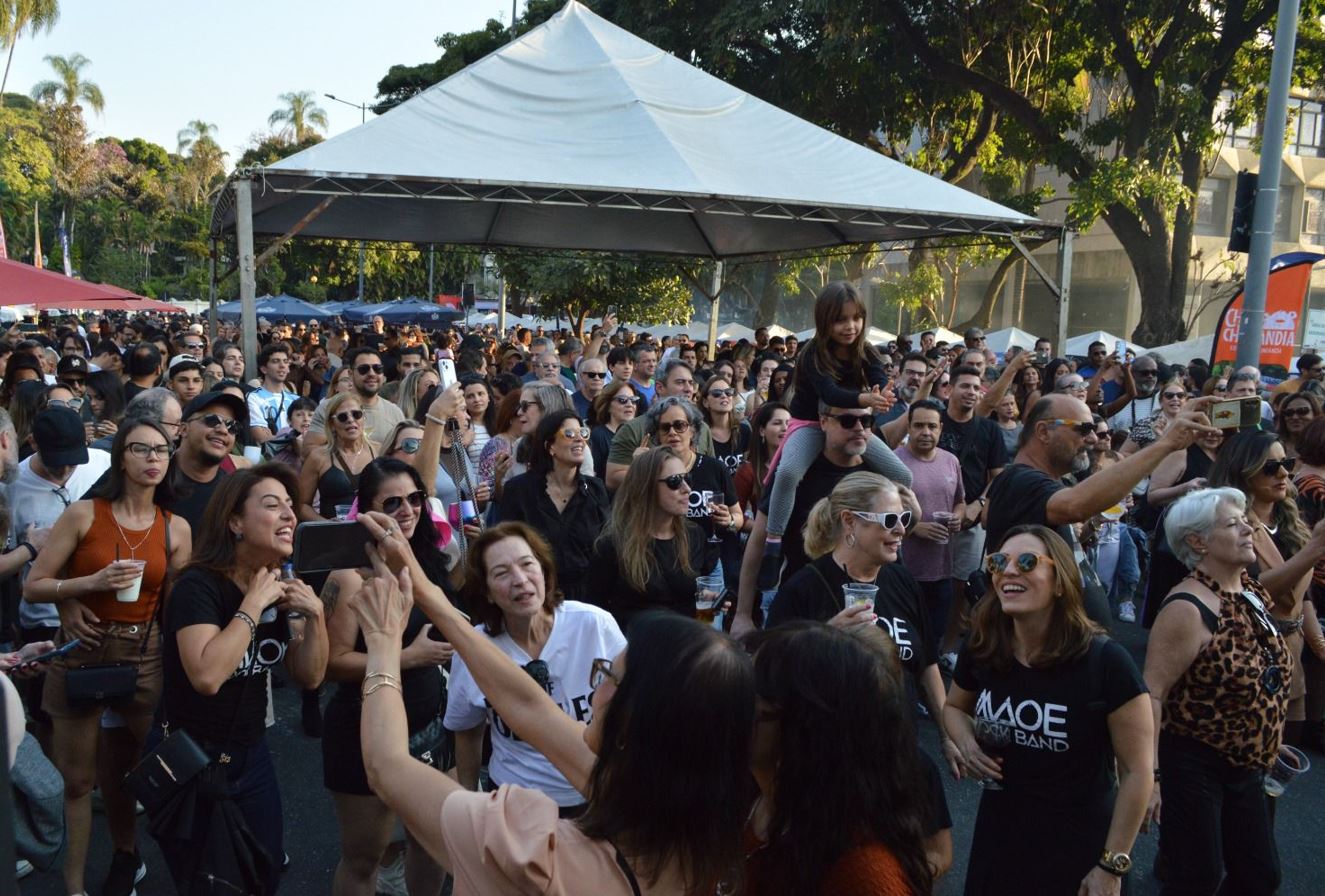 Público numeroso reunido na Praça da Assembleia, em Belo Horizonte, durante evento ao ar livre com palco coberto, pessoas sorrindo, registrando o momento com celulares e aproveitando música, bebidas e clima festivo.