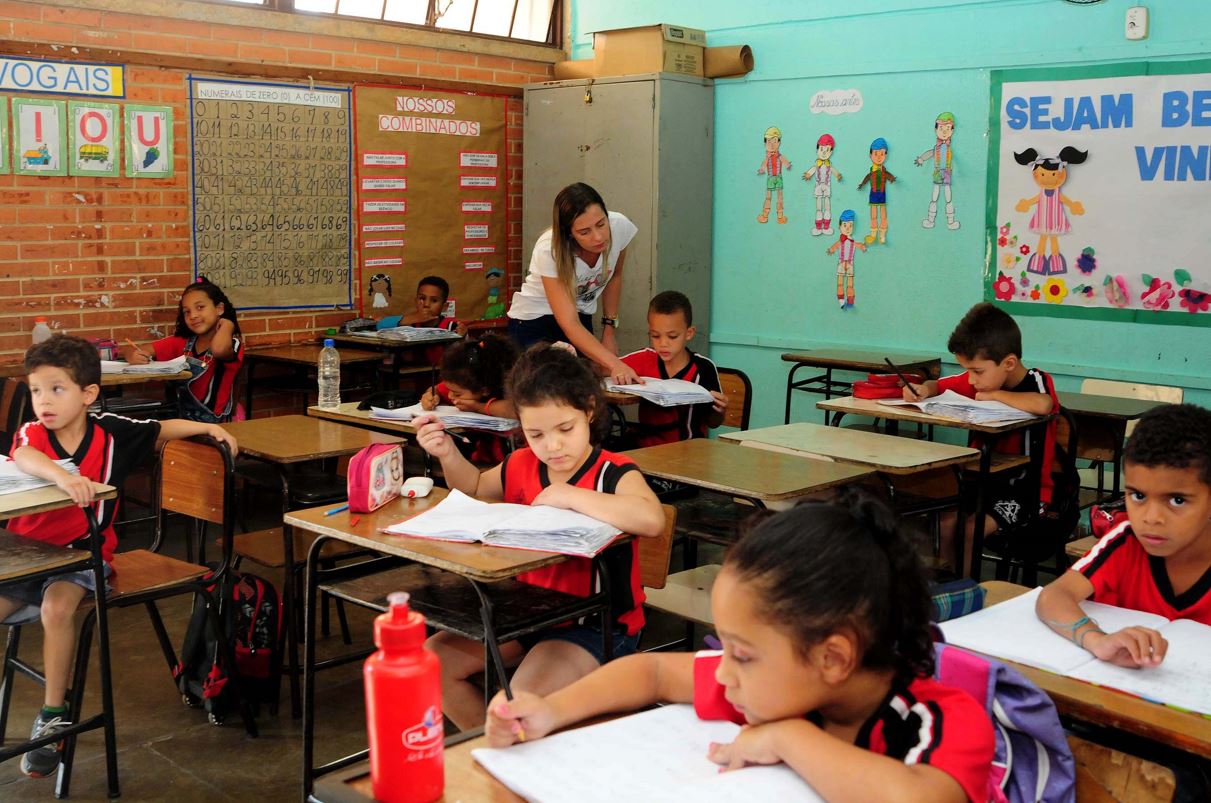 Crianças em sala de aula de escola pública, concentradas em atividades escolares, com professora auxiliando um dos alunos.