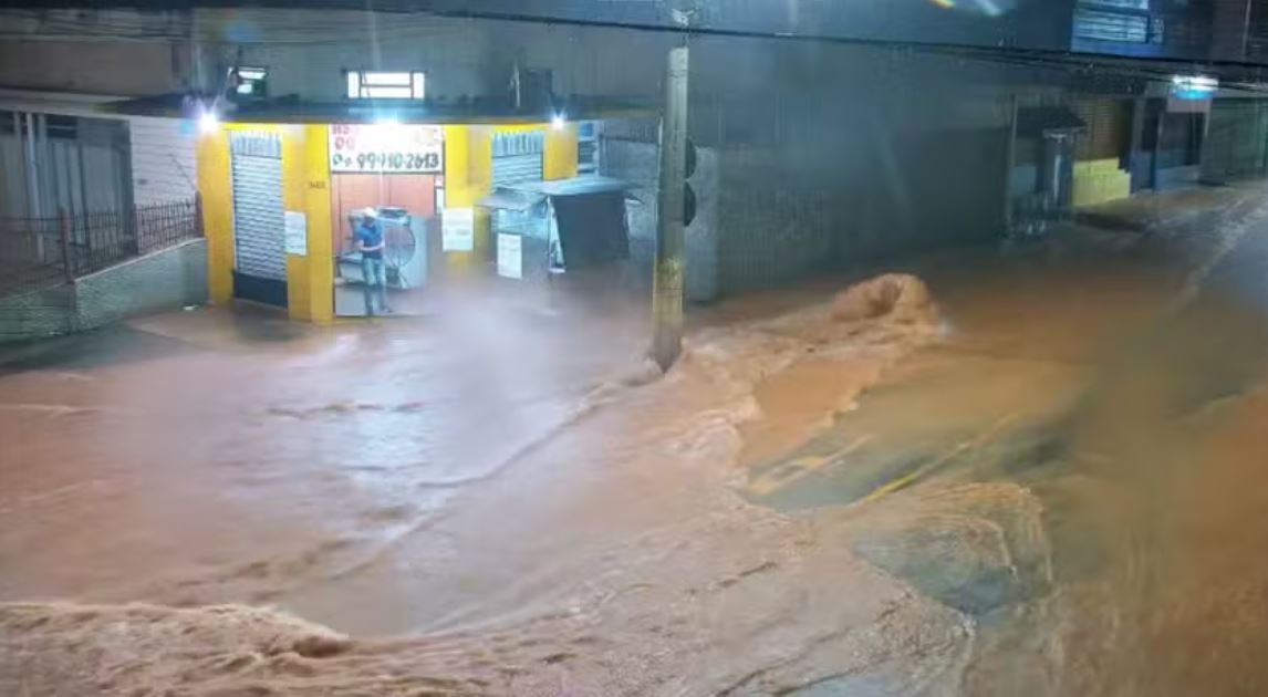 Rua tomada por enxurrada durante temporal em Ubá (MG), com água barrenta invadindo via e estabelecimentos comerciais à noite.