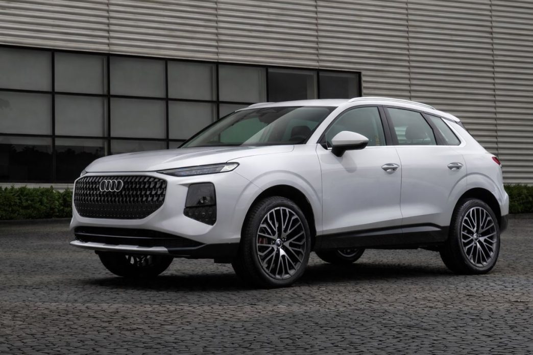 Novo Audi Q3 branco em destaque, visto de frente e lateral, com grade frontal redesenhada e rodas esportivas, estacionado em frente a edifício moderno.