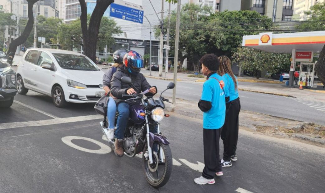 Motociclista com passageira utilizando capacetes realiza transporte por aplicativo em avenida de Belo Horizonte, enquanto dois trabalhadores observam na calçada.
