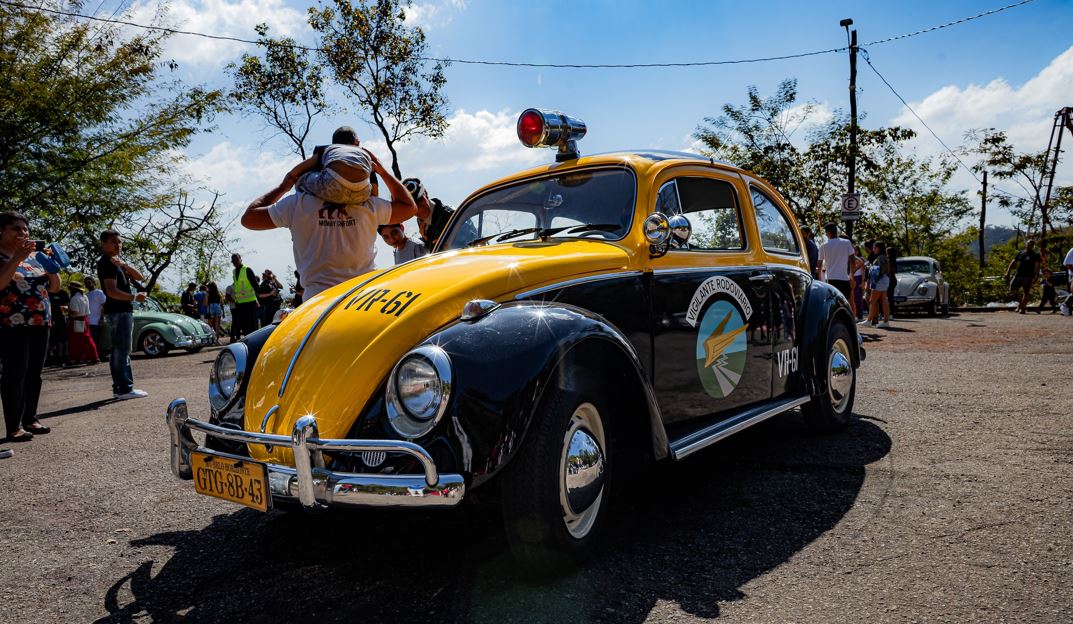 Fusca antigo nas cores amarelo e preto, com sirene no teto, exposto em evento ao ar livre enquanto visitantes circulam e observam outros carros clássicos ao fundo.